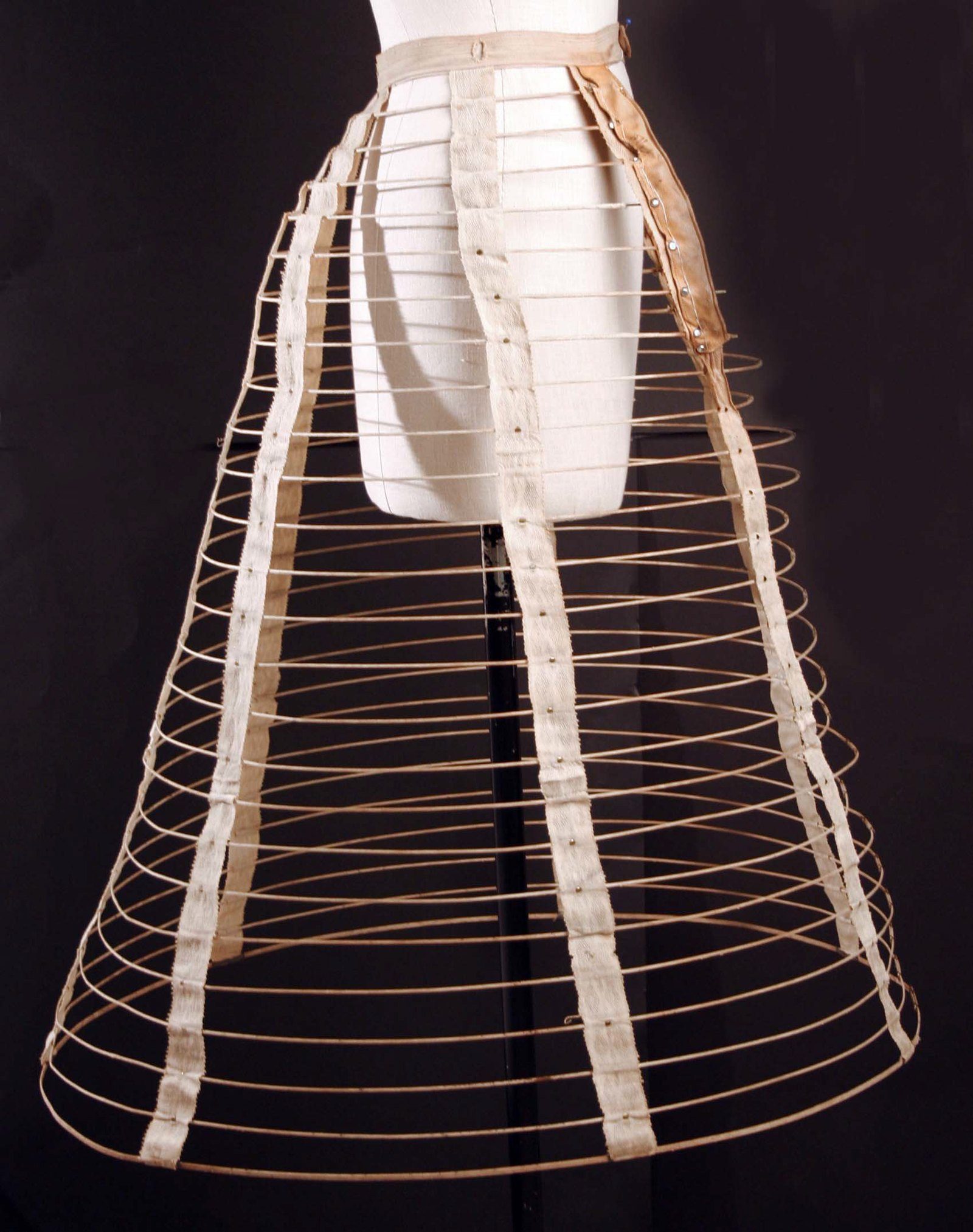 Crinolina de jaula con aros concéntricos y cintas verticales, vista de lado sobre un maniquí
