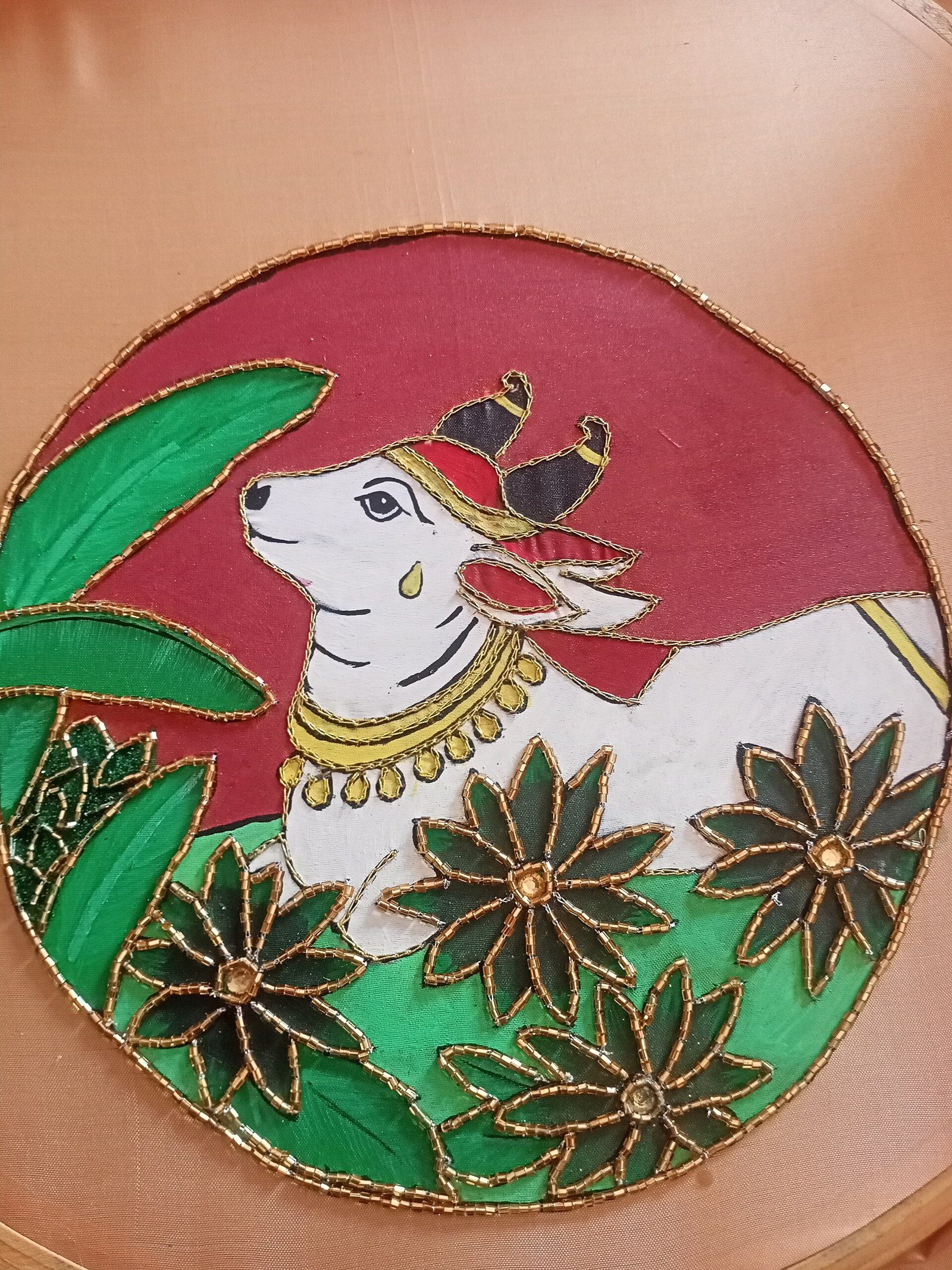 Tela color durazno en bastidor con una vaca blanca y hojas verdes pintadas, bordeadas con cuentas doradas
