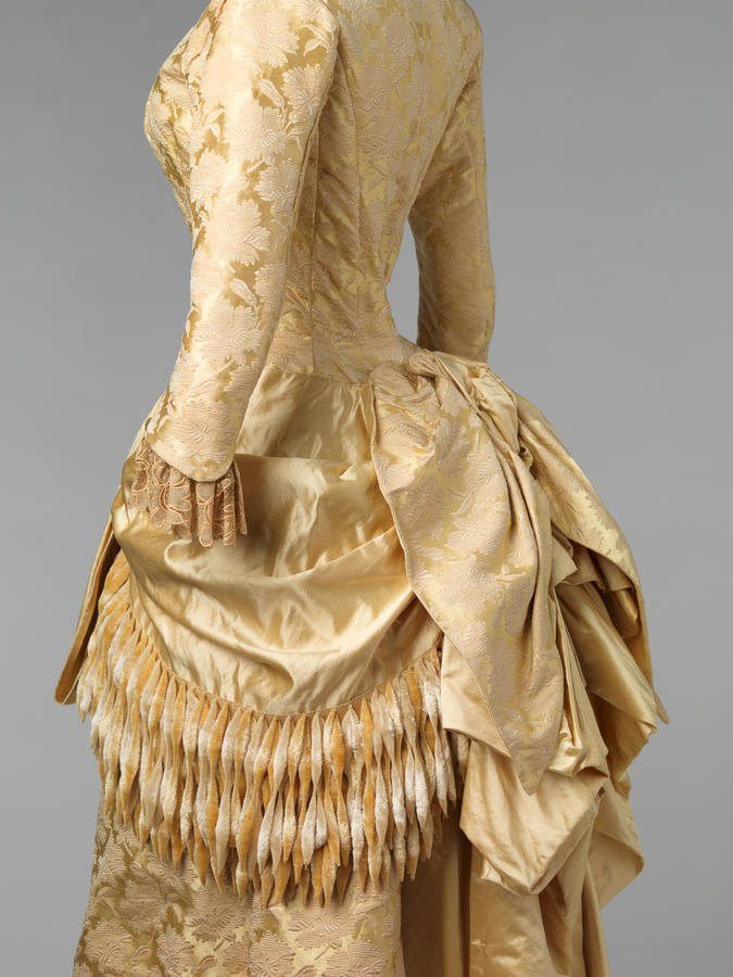 Vestido de recepción de satén y brocado de seda color crema dorado, con drapeado en la falda y flecos de chenilla, sobre fondo gris