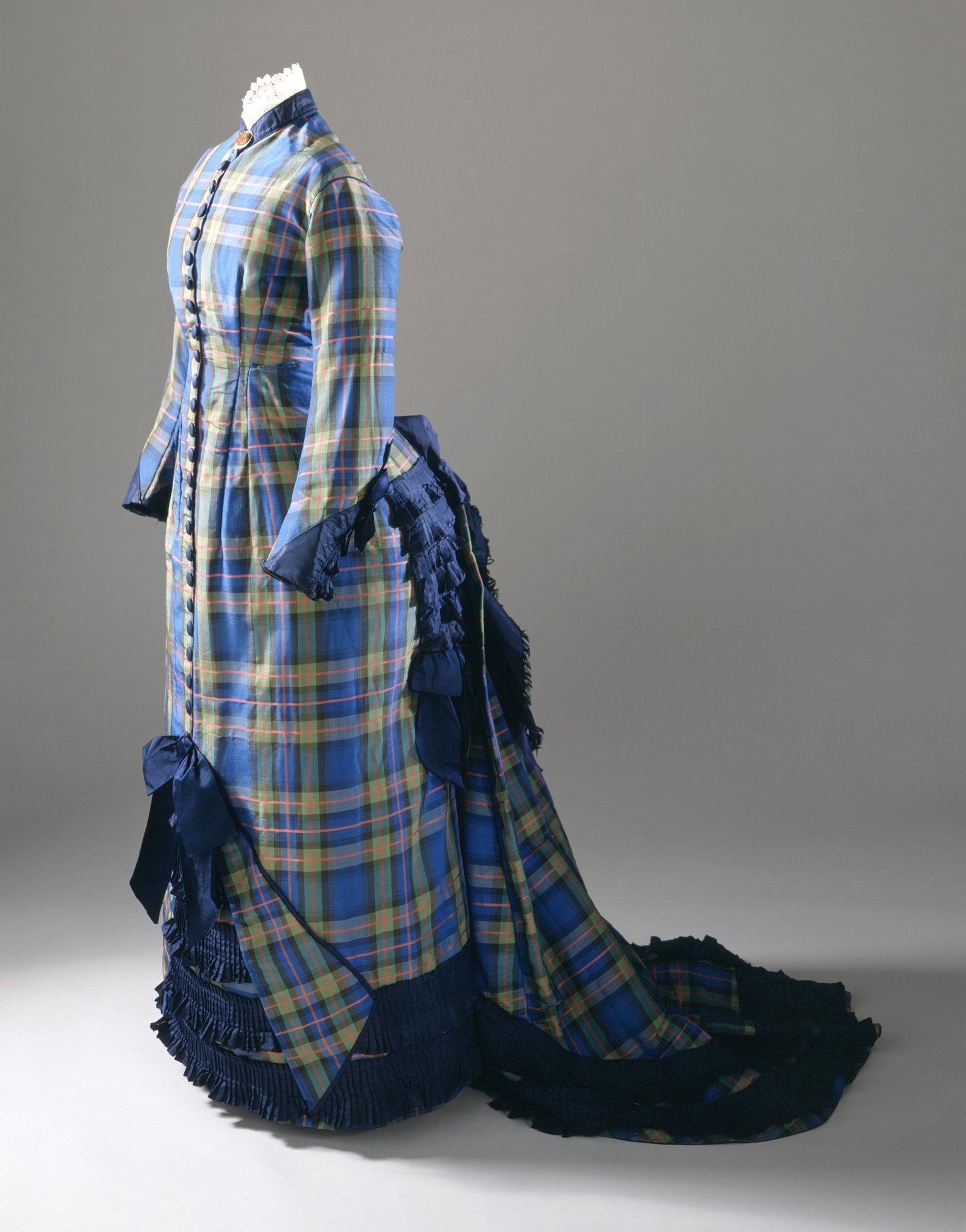 Vestido de seda a cuadros azules y amarillos con botones al frente, cola larga y volantes plisados azul marino en el bajo, sobre un maniquí