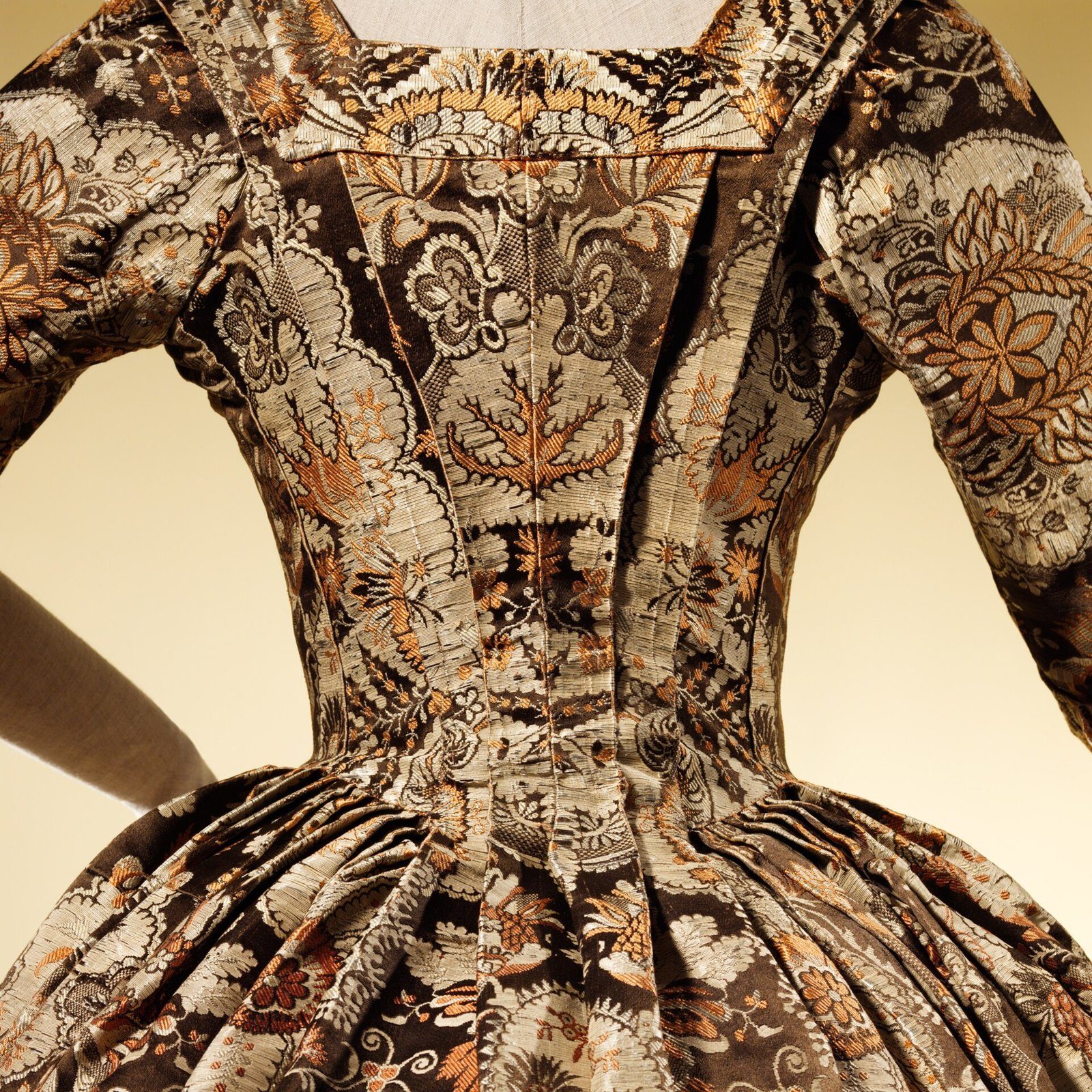 Vestido antiguo de espalda con tela brocada de flores grandes, cuerpo ajustado y falda amplia con pliegues, sobre un maniquí gris